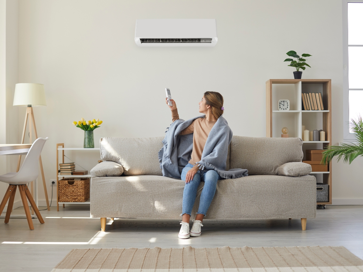 A woman sits on a sofa with a blanket, using a remote to operate a wall-mounted air conditioner in her bright, modern living room, enjoying improved indoor air quality Atlanta residents appreciate.