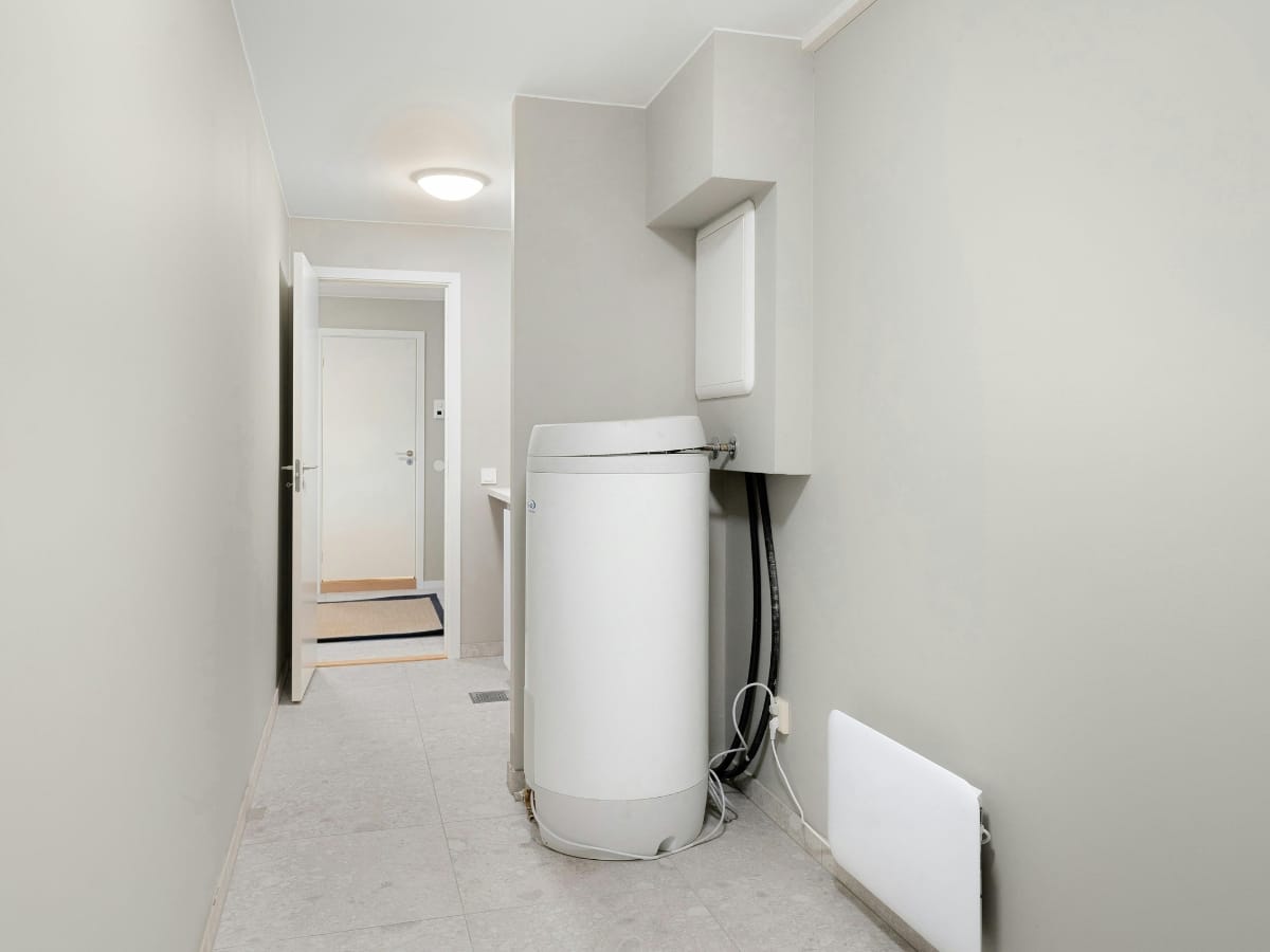 A white washing machine sits against a light gray wall in a narrow hallway with tiled flooring, near a door leading to an adjacent room—perfectly complementing homes equipped with modern tankless water heaters.