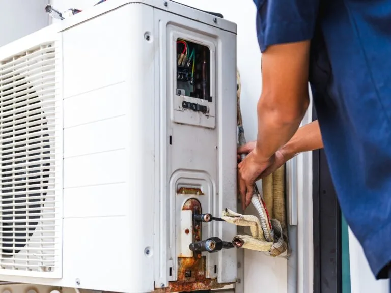 A person is repairing or inspecting an outdoor air conditioning unit with tools and exposed wiring visible, ensuring optimal HVAC installation timing for efficient cooling.