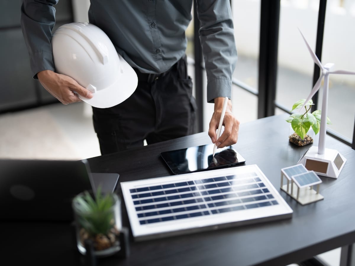 Person stands at a desk holding a white hard hat, with a tablet, laptop, solar panel, and wind turbine models displayed on the table—showcasing smart ways to reduce energy bills with innovative technology.