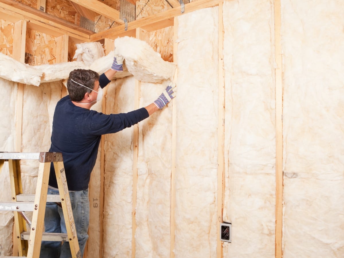 A man wearing gloves and a mask installs fiberglass insulation in the wall of a wooden-framed building under construction to help reduce energy bills.