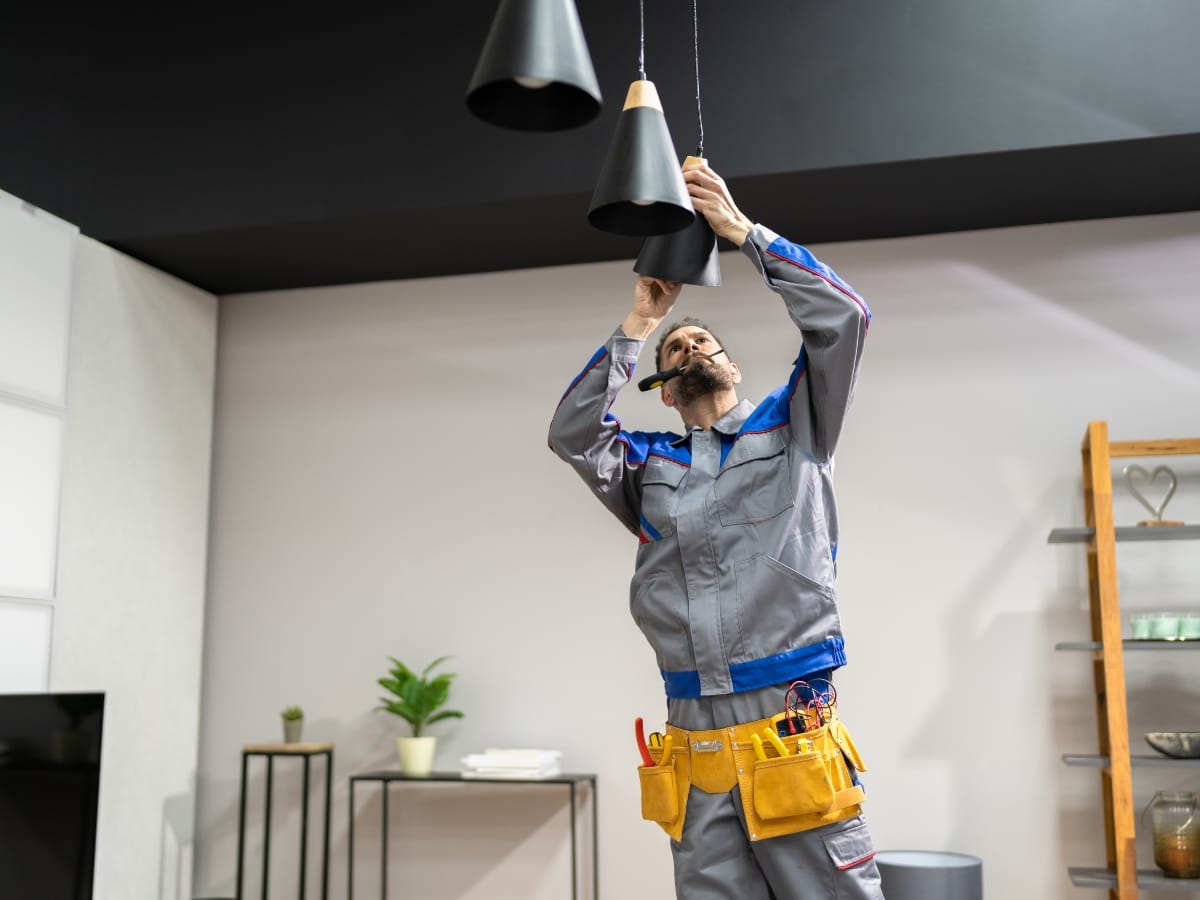 Electrician in a gray uniform stands on a step stool and adjusts a ceiling light fixture in a modern room, performing tasks from a home maintenance checklist Georgia residents can rely on.