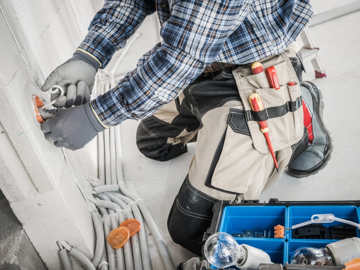 Electrician wearing gloves installs or repairs an electrical outlet; various tools and cables are visible, along with an open toolbox on the floor—ideal for your home maintenance checklist in Georgia.