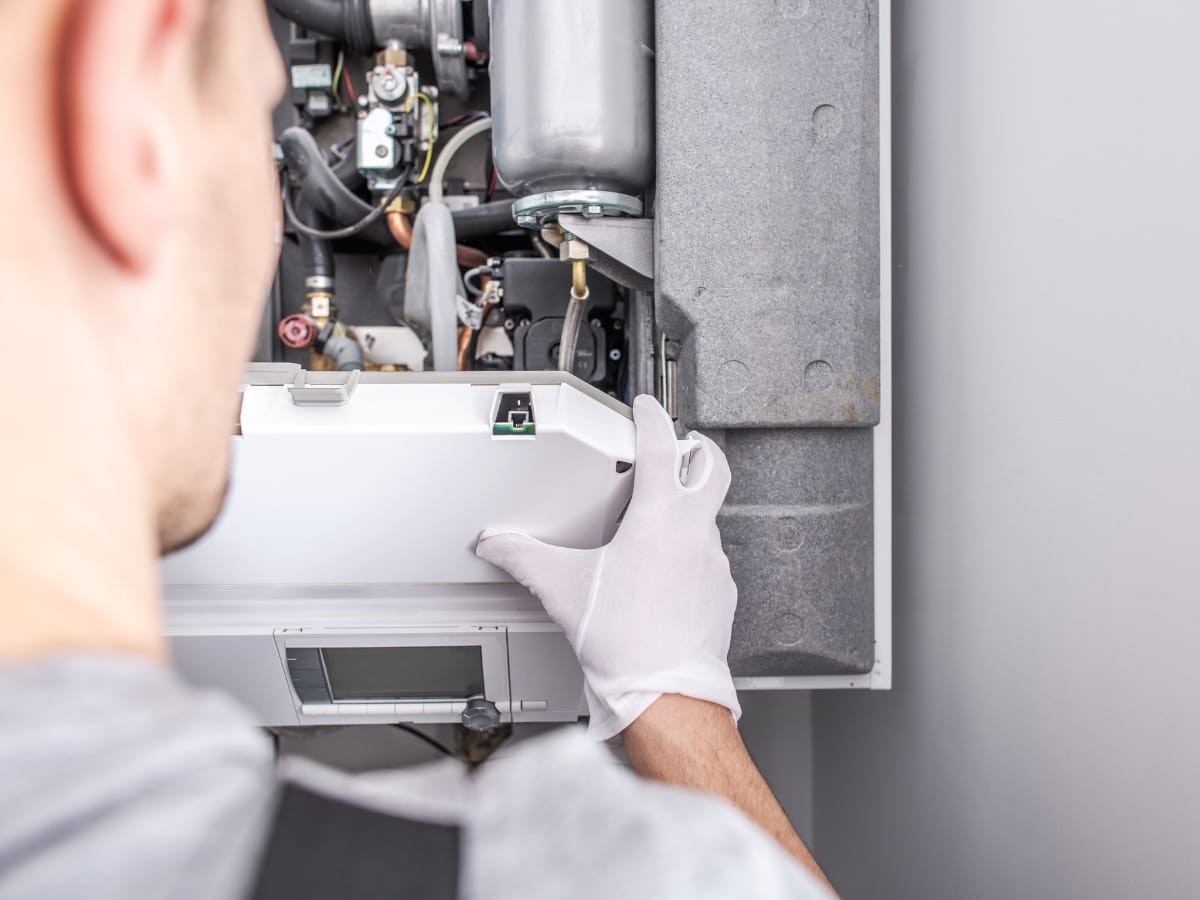A person wearing white gloves inspects or repairs the internal components of a wall-mounted HVAC or boiler system as part of a home maintenance checklist Georgia homeowners can follow.