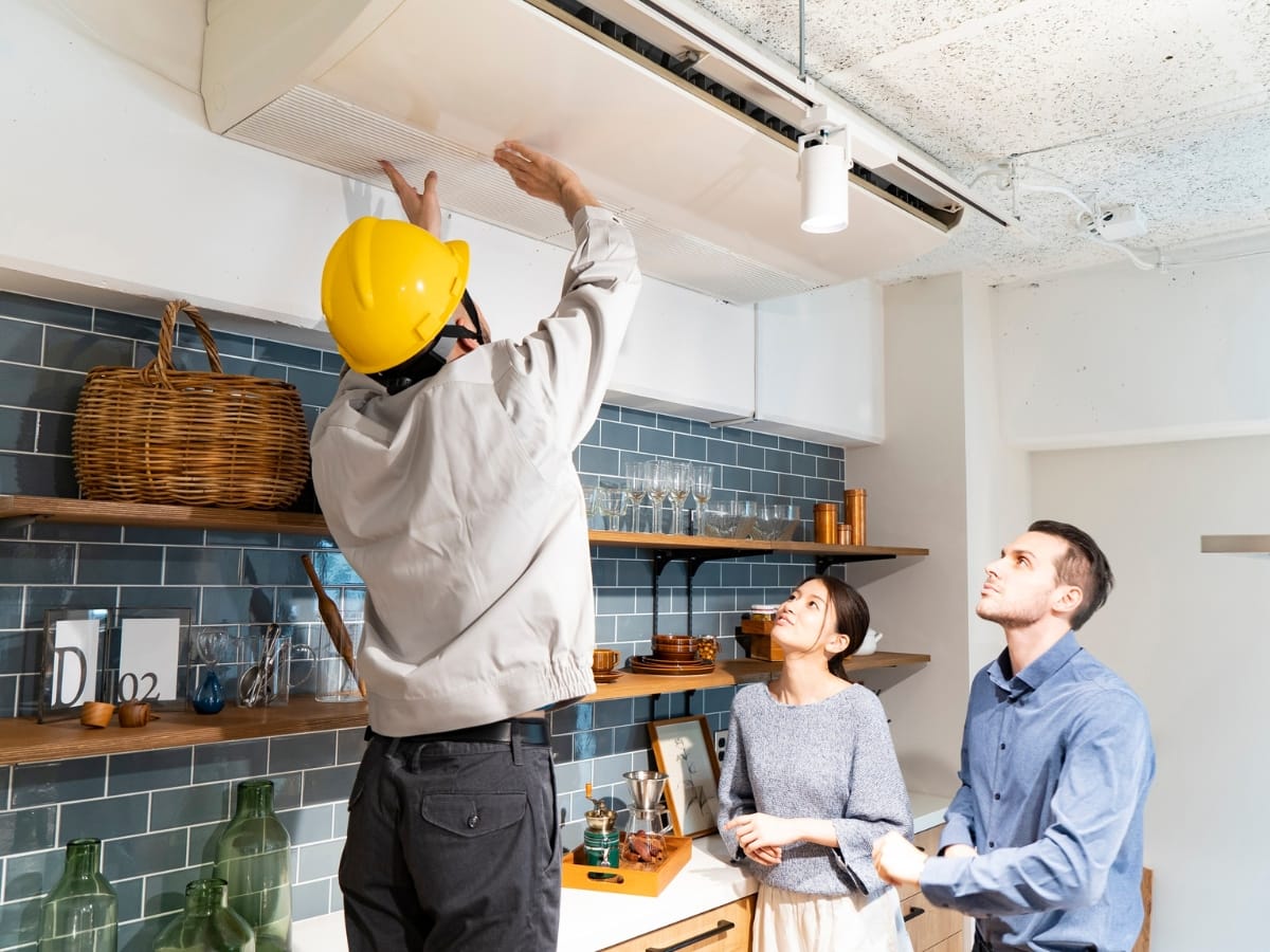 A worker in a yellow helmet repairs an air conditioning unit while two people watch in a modern kitchen, highlighting the importance of following a home maintenance checklist Georgia residents recommend.