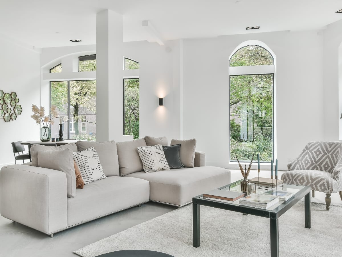 Modern living room with a light gray sectional sofa, patterned armchair, glass coffee table, and large windows letting in natural light—showcasing popular home renovation trends Atlanta homeowners love, with views of greenery outside.