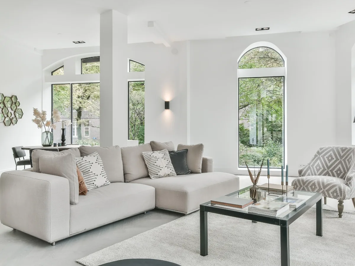 Modern living room with a light gray sectional sofa, patterned armchair, glass coffee table, and large windows letting in natural light—showcasing popular home renovation trends Atlanta homeowners love, with views of greenery outside.