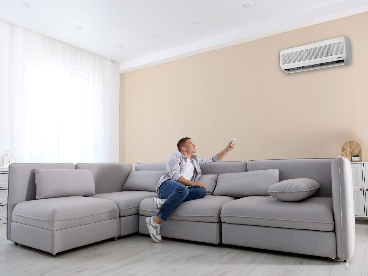 A man sitting on a gray sectional sofa points a remote control at a wall-mounted, allergy friendly HVAC unit in a bright living room.