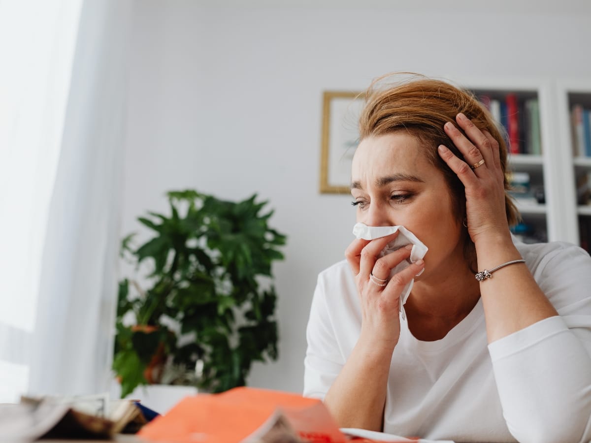 Woman in a white shirt sits at a table holding a tissue to her nose, appearing unwell—possibly feeling the humidity health impact—with one hand on her head. Indoor setting with plants and bookshelves in the background.