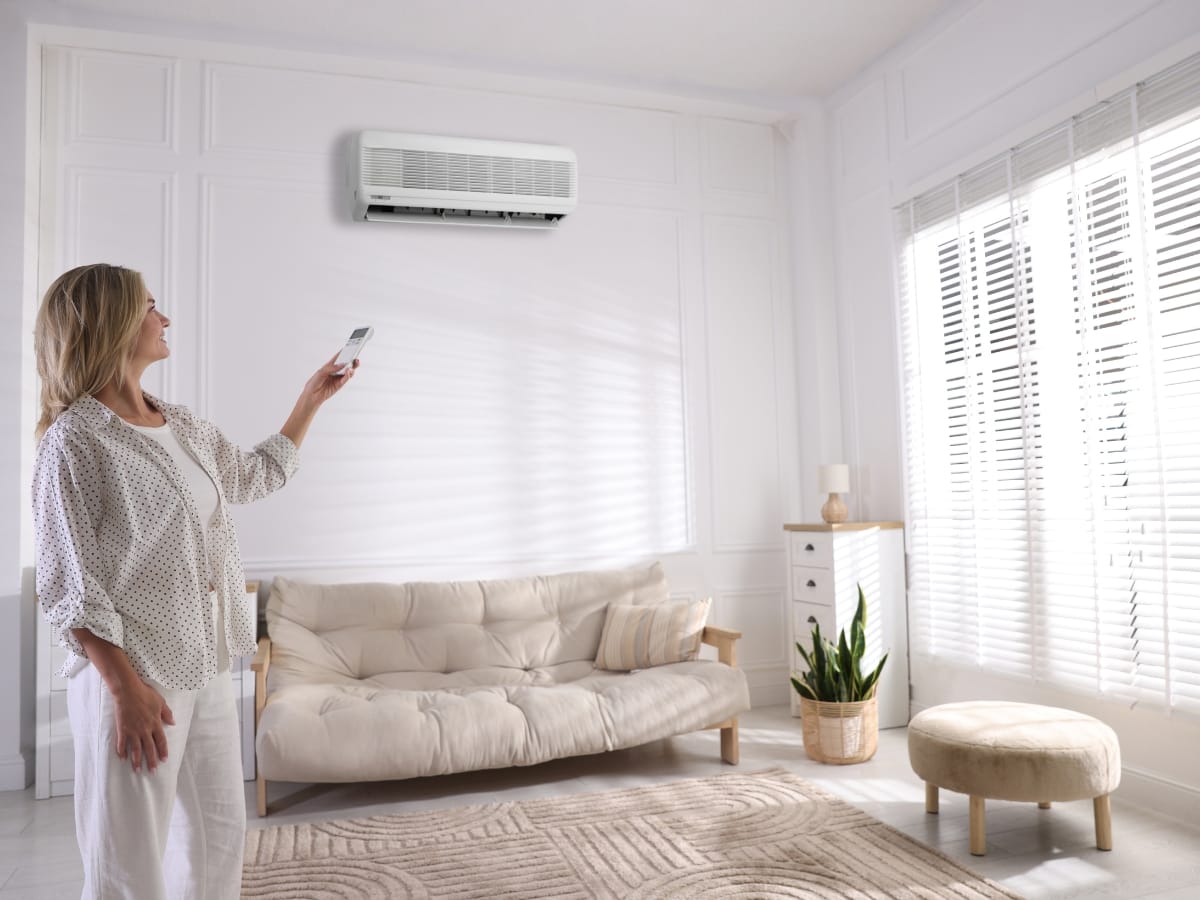 A woman stands in a bright living room using a remote control to operate a wall-mounted air conditioner, helping to manage indoor comfort and minimize the humidity health impact.