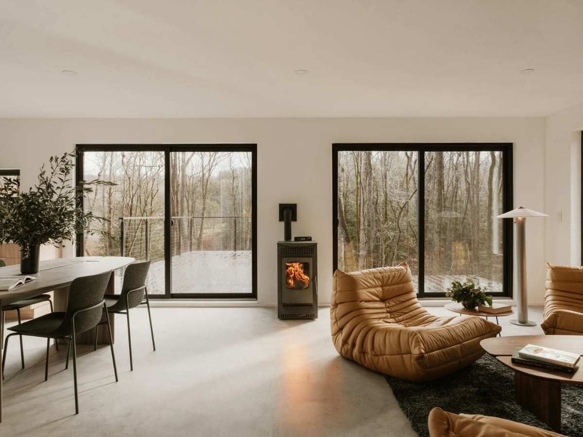 A modern living room with large windows, tan lounge chairs, a wood-burning stove, and a dining table with black chairs overlooks a forested outdoor deck, designed to optimize views and minimize the humidity health impact.