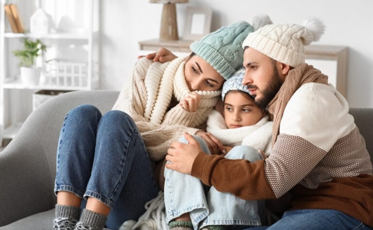 A family of three sits close together on a couch, all wearing winter hats, scarves, and sweaters, looking pensive and thoughtful—perhaps pondering some much-needed furnace troubleshooting.