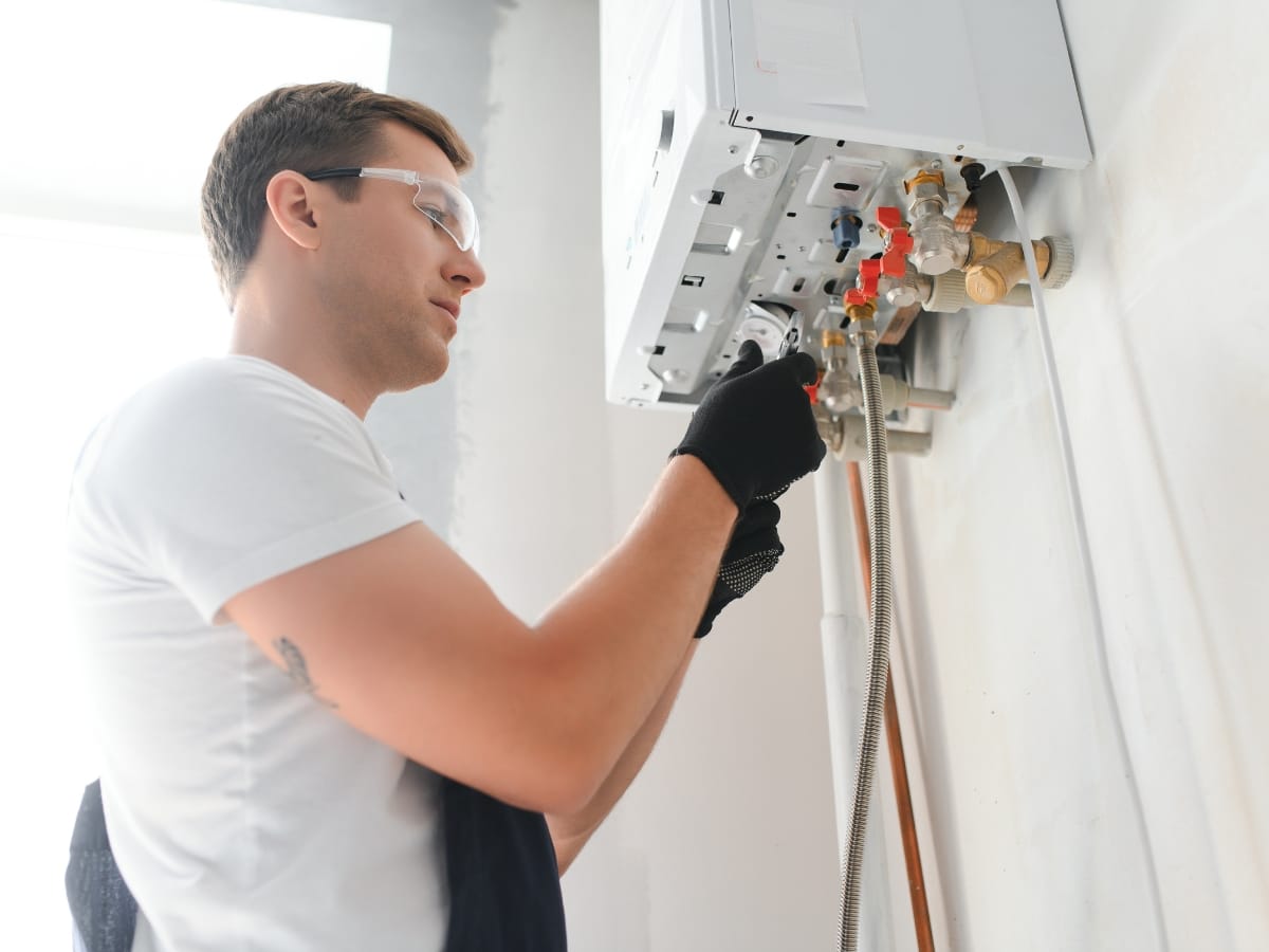 A person wearing safety glasses and gloves is repairing or inspecting a wall-mounted gas boiler with exposed pipes, possibly checking the furnace pilot light to ensure safe operation.