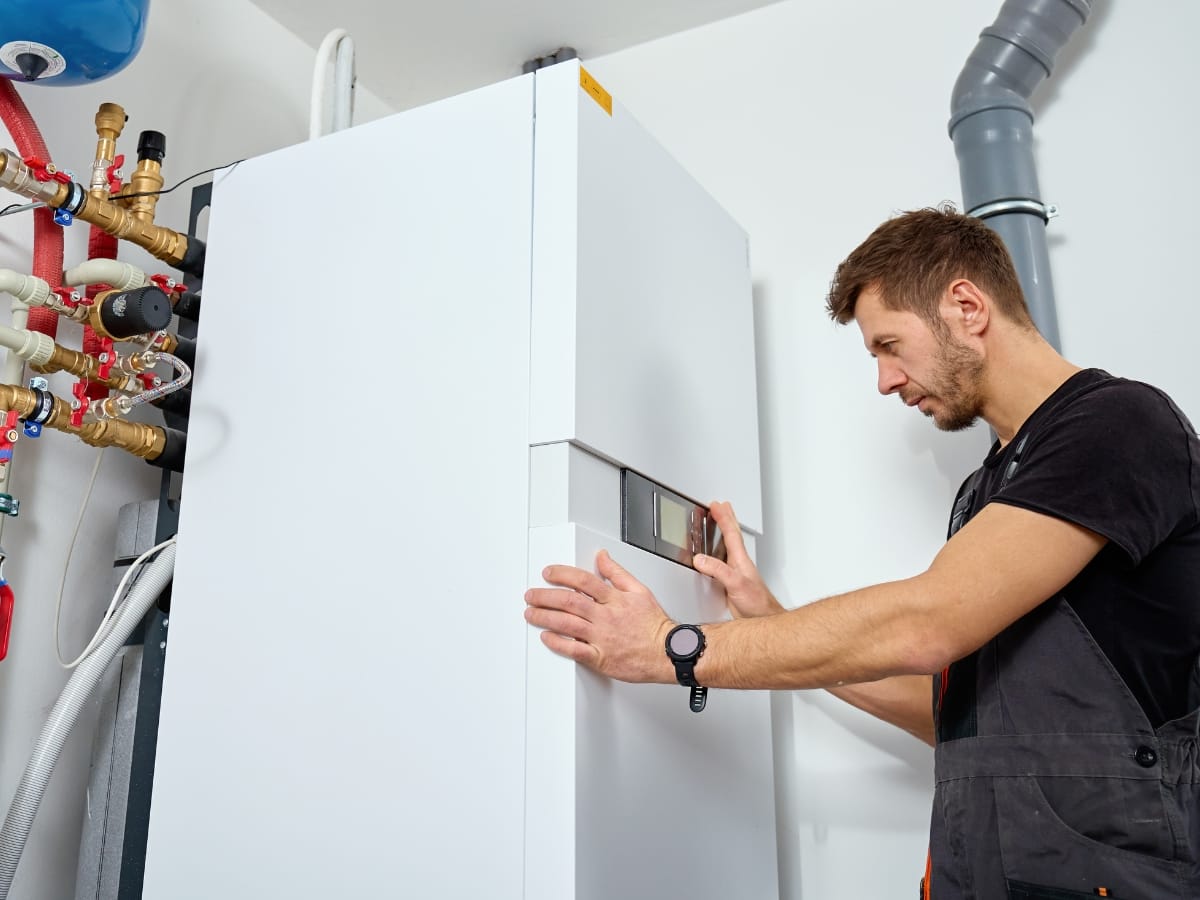 A technician adjusts the settings on a white boiler unit in a utility room, carefully checking the furnace pilot light among various pipes and valves.
