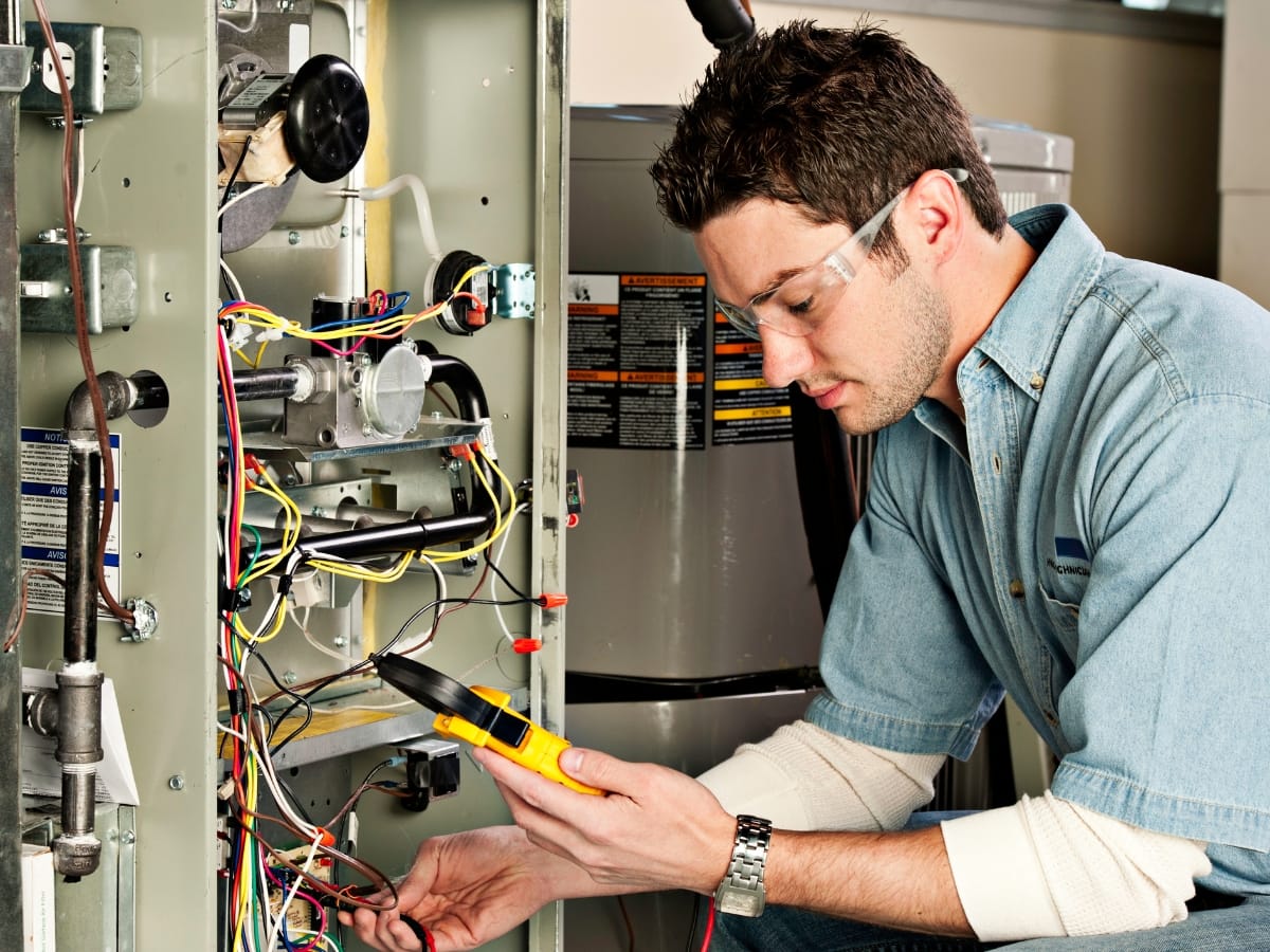 A technician wearing safety glasses uses a multimeter to check electrical connections inside an open HVAC unit during furnace troubleshooting.