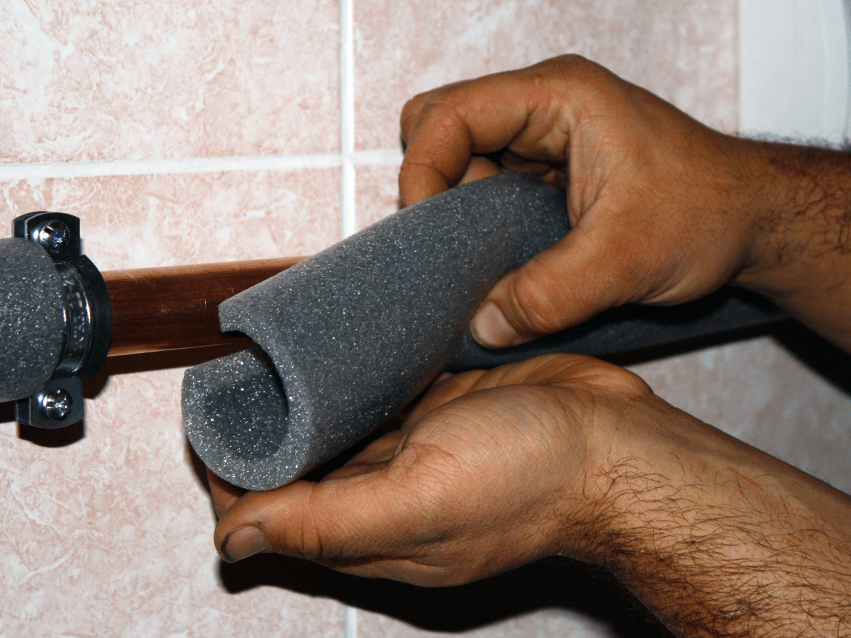 Hands installing foam pipe insulation on a copper pipe against a tiled wall to help prevent burst pipes.
