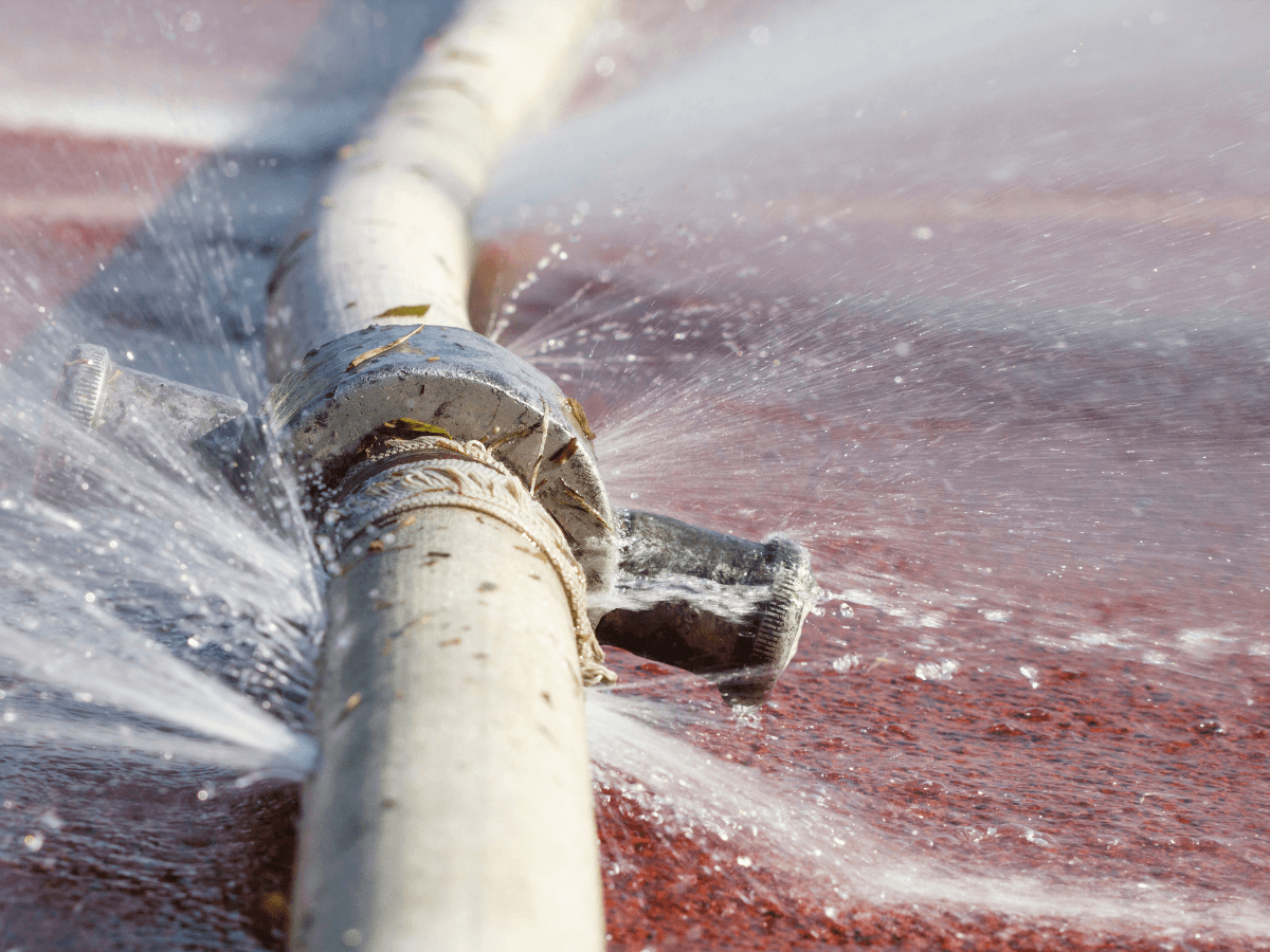 A metal pipe with a leaking joint sprays water in multiple directions onto a red surface, highlighting the importance of maintenance to prevent burst pipes.