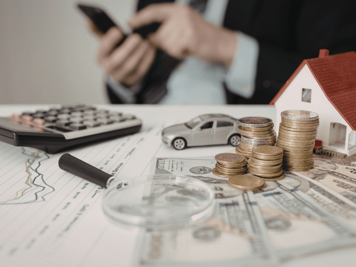 A person using a smartphone sits behind stacks of coins, a model house, toy car, calculator, magnifying glass, and US dollar bills on financial documents—perhaps researching ways to save money and prevent burst pipes in their home.