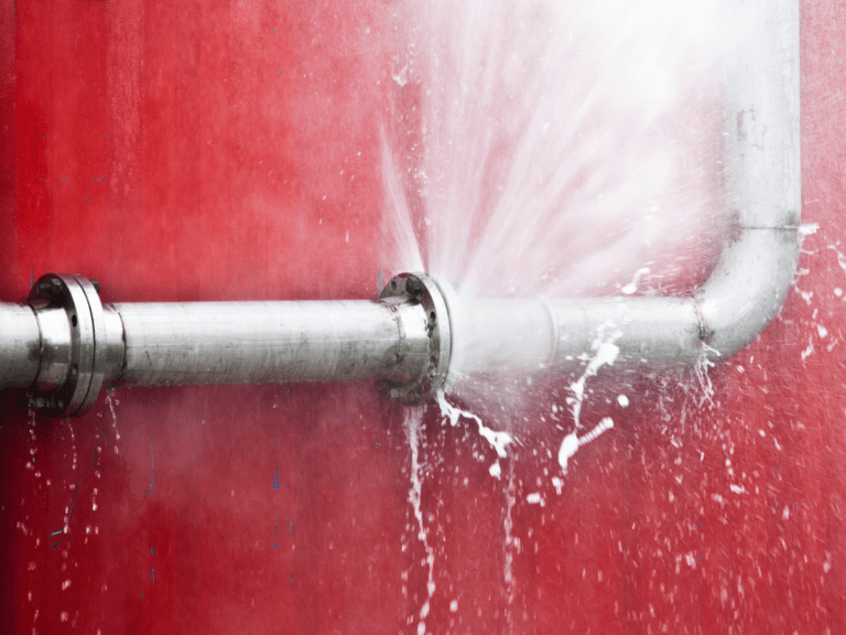 A burst metal pipe sprays water forcefully against a red wall—highlighting the urgent need to prevent burst pipes before costly damage occurs.