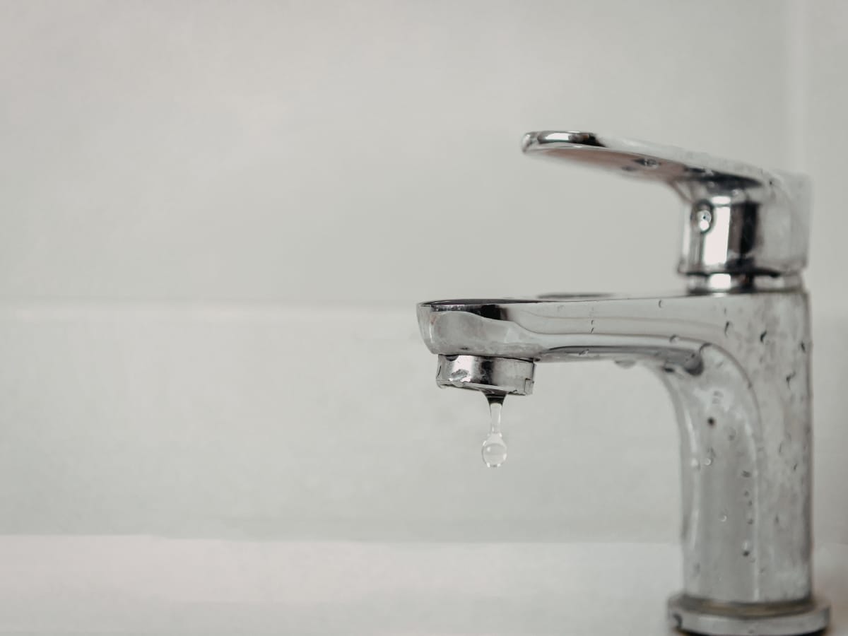 A close-up of a chrome bathroom faucet captures a single droplet of water falling from the spout, highlighting the need for expert Faucet Repair Installation against a plain light background.