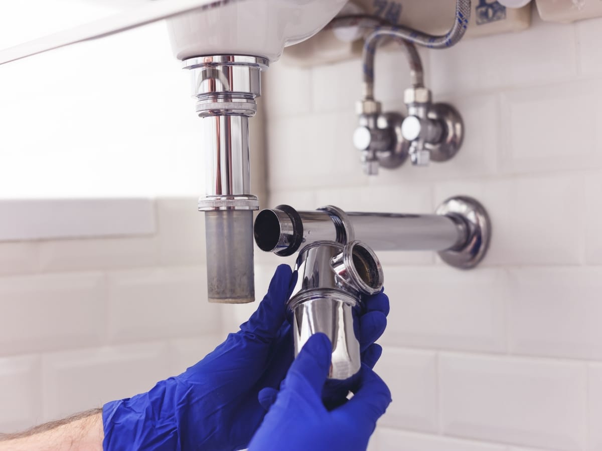 Person wearing blue gloves removes a chrome P-trap pipe under a sink for plumbing maintenance and faucet repair installation against a white tiled wall.