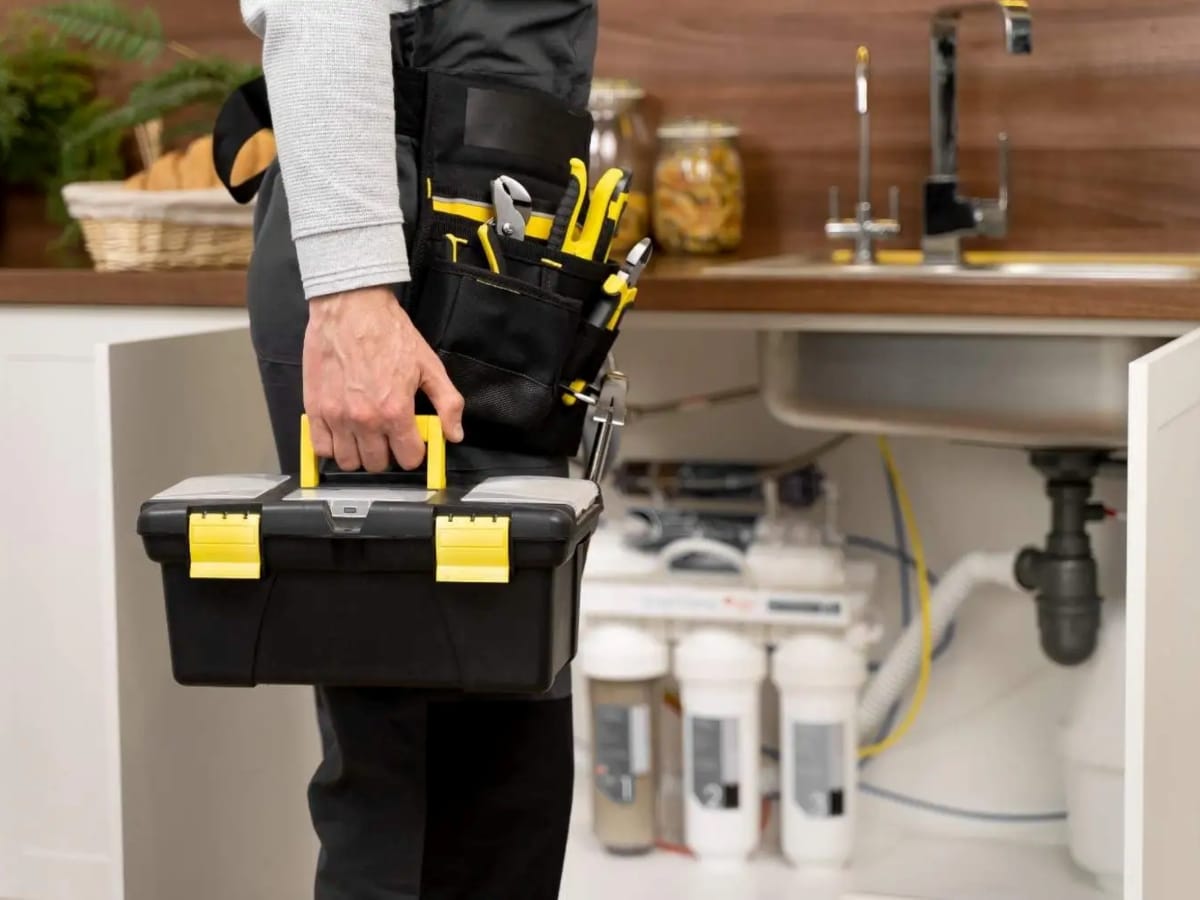 Person holding a toolbox and standing by a kitchen sink cabinet, wearing a tool belt with various tools; water filtration system and pipes visible under the sink, ready for faucet repair installation.