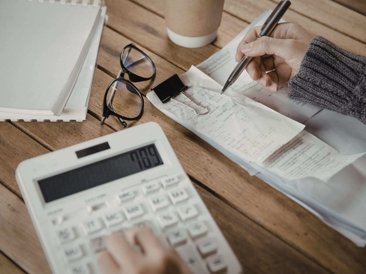 Person using a calculator and pen to review receipts at a wooden table, possibly organizing expenses after a recent faucet repair installation, with a notebook, eyeglasses, and coffee cup nearby.