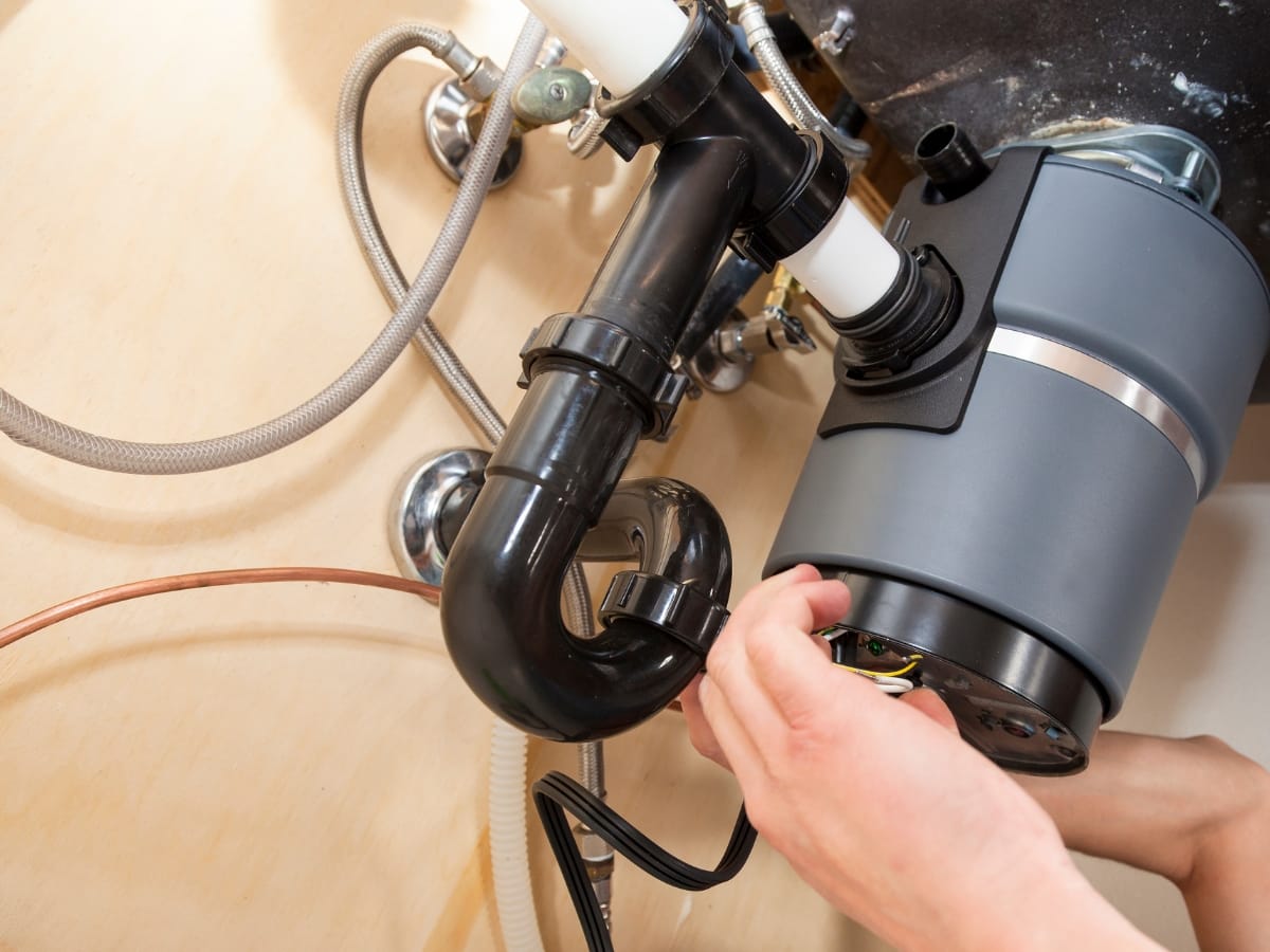 Person performing garbage disposal repair under a kitchen sink, working on visible pipes and connectors.