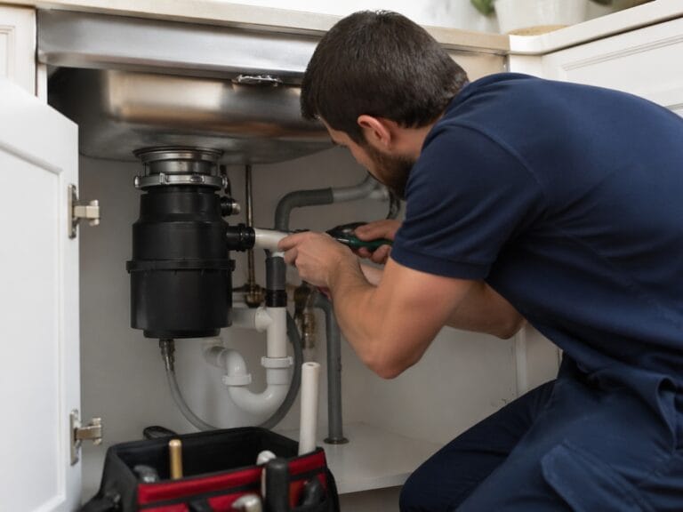 A plumber in a navy shirt uses a screwdriver to repair pipes and perform garbage disposal repair under a kitchen sink.