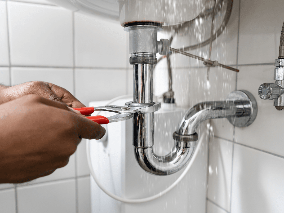 A person uses pliers to fix a leaking pipe under a sink, with water spraying out in a tiled bathroom—while smart leak detectors alert them to the issue.