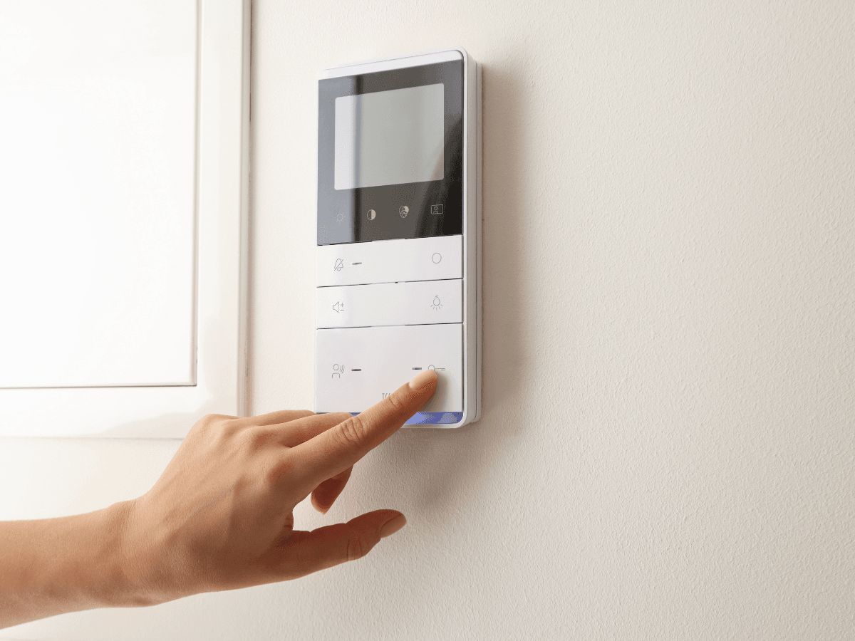 A hand presses a button on a wall-mounted touch screen control panel with a blank display, next to a white wall and window—perfect for learning thermostat programming tips.