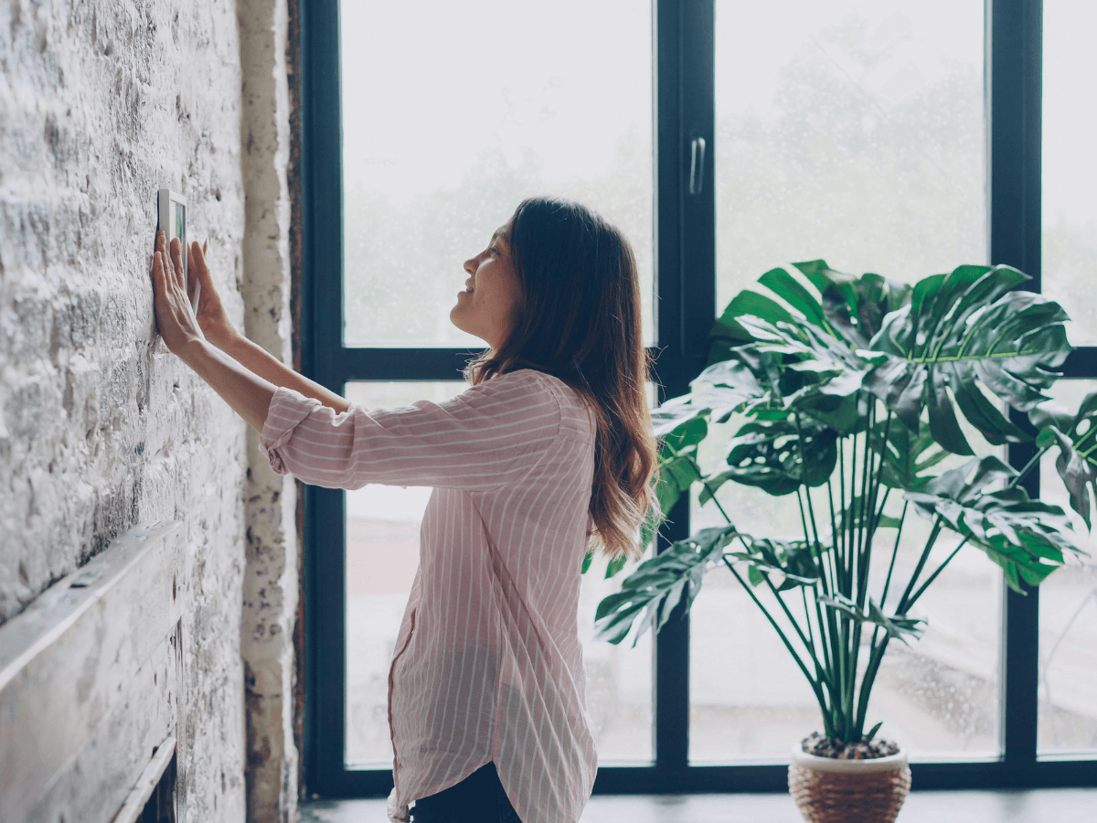 Woman adjusts a thermostat on a brick wall in a room with large windows and a potted green plant, showcasing smart thermostat programming tips for comfort and energy savings.