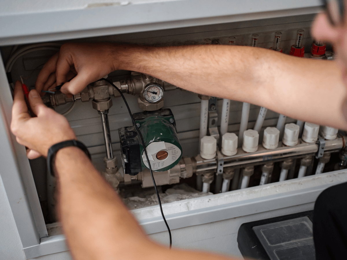A person adjusts wires on a control panel with valves, a pressure gauge, and a pump, likely performing HVAC maintenance or repairs to ensure efficient operation and reduce climate impact.