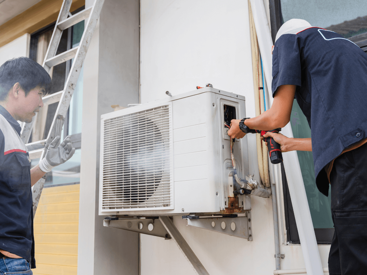 Two technicians are servicing an outdoor air conditioning unit mounted on a building wall, mindful of climate impact; one uses tools while the other stands nearby.