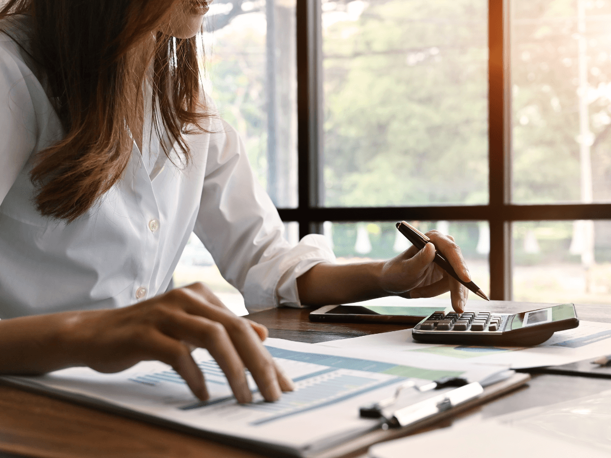 A person in a white shirt uses a calculator and reviews financial documents at a desk near large windows with sunlight, assessing the climate impact of HVAC systems.