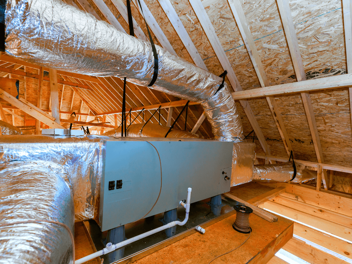 An HVAC air handler unit with insulated ducts, designed for reduced climate impact, is installed in the attic of a house under construction, surrounded by exposed wooden framing and roof trusses.