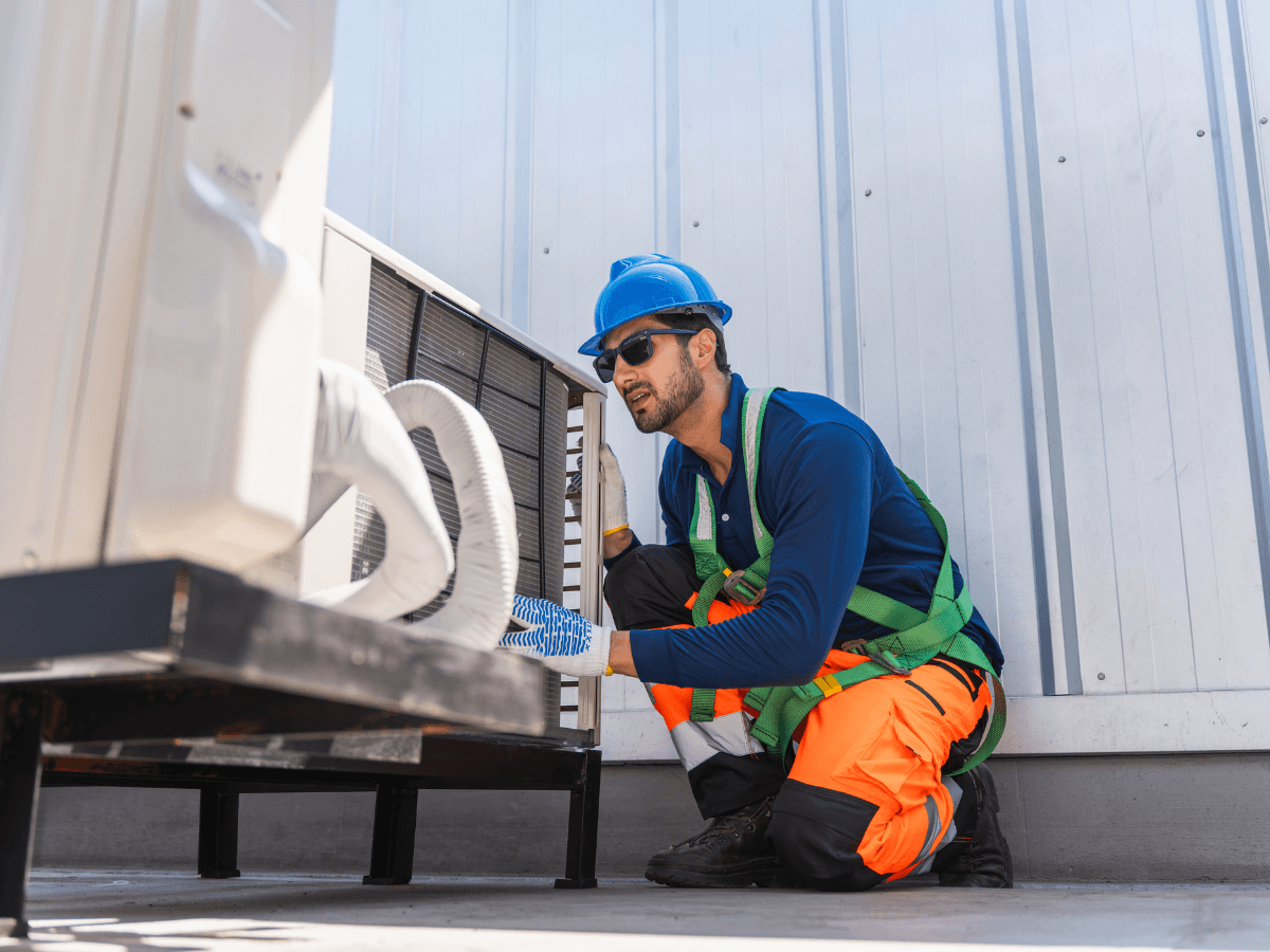 A worker in safety gear kneels to inspect or repair an air conditioning unit on a rooftop next to a metal building wall, ensuring the HVAC system operates efficiently to help maintain home value.