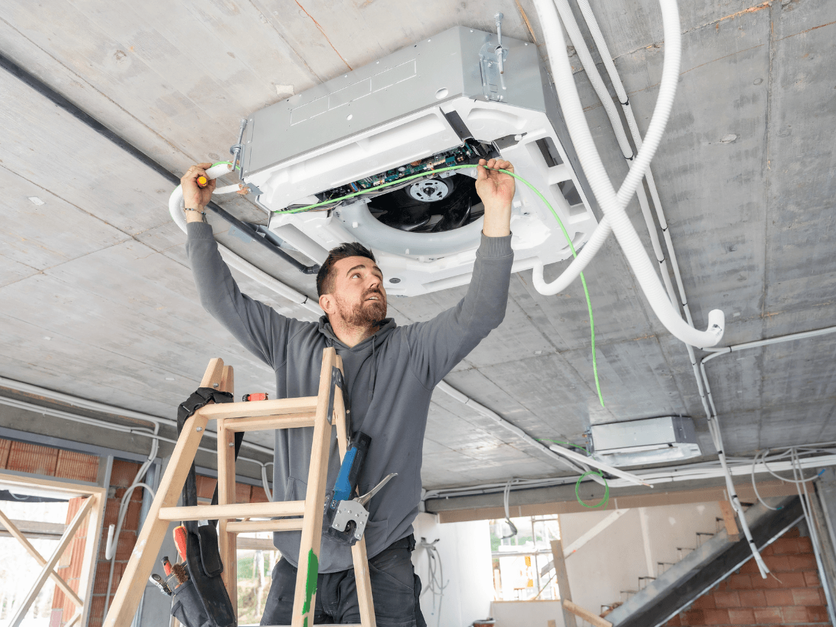 A man standing on a ladder installs or repairs a ceiling-mounted air conditioning unit in a building under construction, enhancing home value with efficient HVAC solutions. Various tools and wires are visible around the work area.