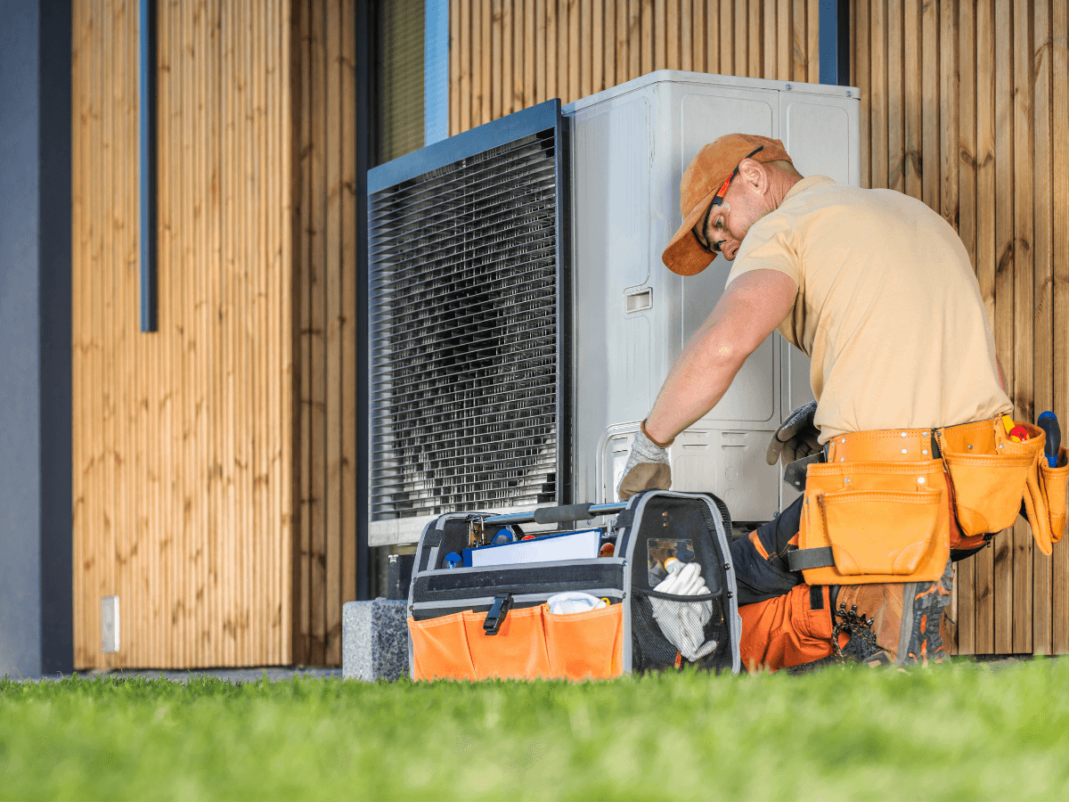A technician in an orange cap and tool belt services an outdoor air conditioning unit next to a modern building, with tools organized in a bag on the ground—demonstrating how regular HVAC maintenance can help protect your home value.
