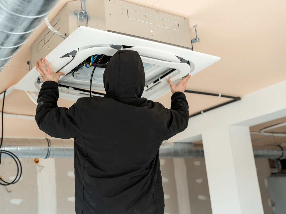 Person in a black hoodie installs or repairs a ceiling-mounted HVAC unit in a room under construction, ensuring optimal climate control and enhancing home value.