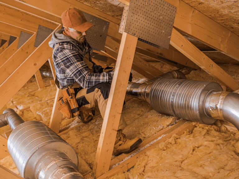 A worker in a flannel shirt and cap is installing or inspecting metal air ducts in an unfinished attic with exposed insulation and wooden beams, ensuring the HVAC system operates efficiently to help boost home value.