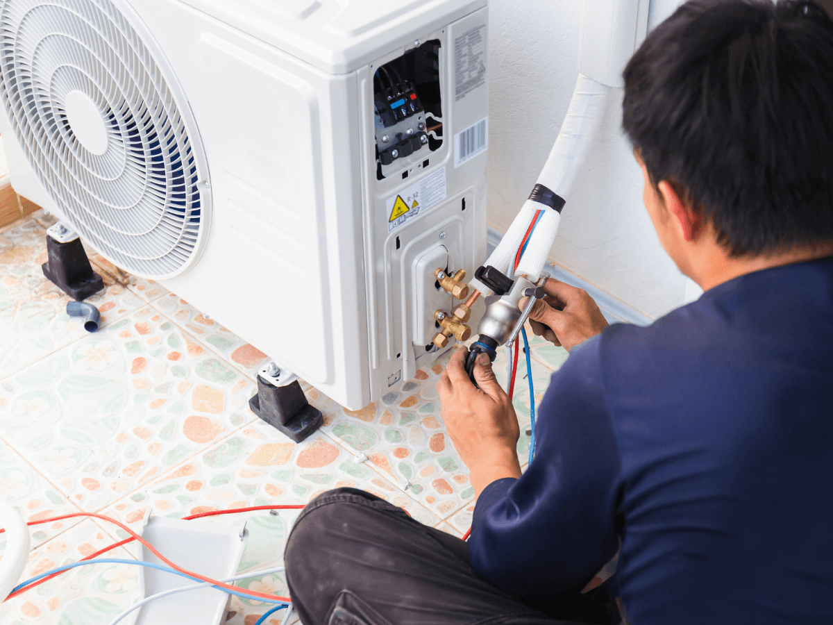 A technician installs or repairs the outdoor unit of an air conditioning system, connecting pipes and wiring on a tiled floor, while performing an AC refrigerant recharge to ensure optimal cooling performance.
