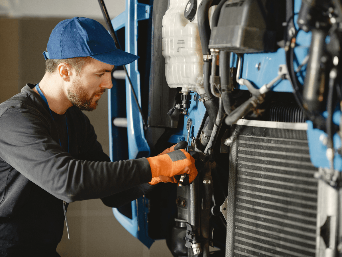 Mechanic wearing a blue cap and orange gloves is performing an AC refrigerant recharge while working on the engine of a blue vehicle inside a garage.