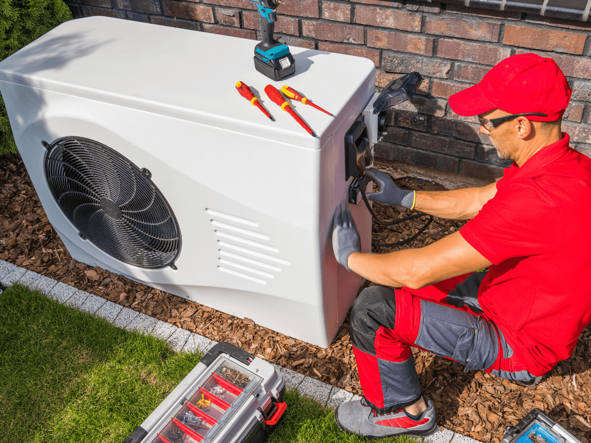 Technician in red uniform servicing an outdoor heat pump unit with tools and equipment on the ground nearby, performing an AC refrigerant recharge.