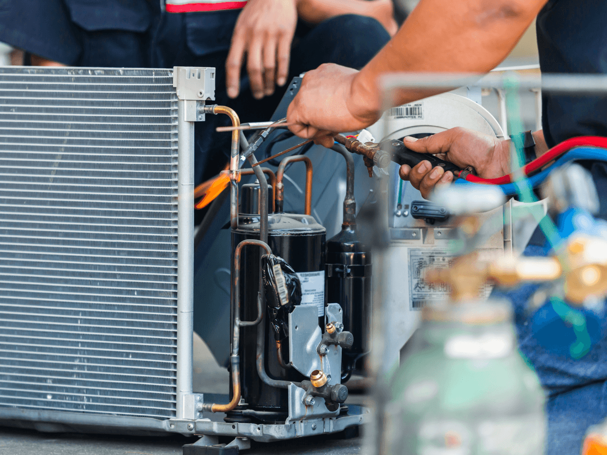 A technician uses tools to repair or install parts on an air conditioning unit, possibly performing an AC refrigerant recharge, with another person visible in the background.
