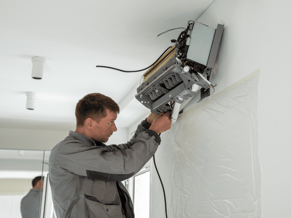 A technician wearing a gray jacket performs ductless mini-split repair, working on an air conditioning unit mounted high on a white wall in a modern room.