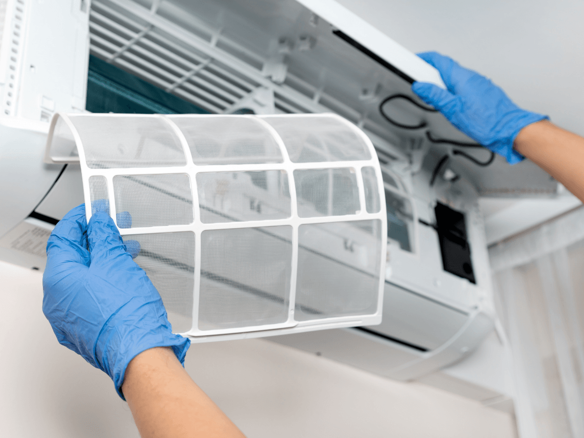 Person wearing blue gloves removes a mesh filter from a wall-mounted air conditioner for cleaning or maintenance, demonstrating an important step in ductless mini-split repair.