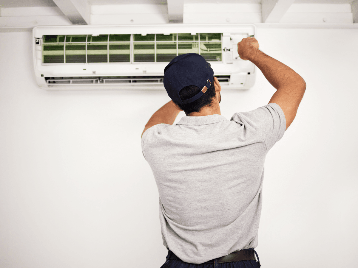 A person in a gray shirt and cap is servicing or repairing a wall-mounted ductless mini-split air conditioner indoors.