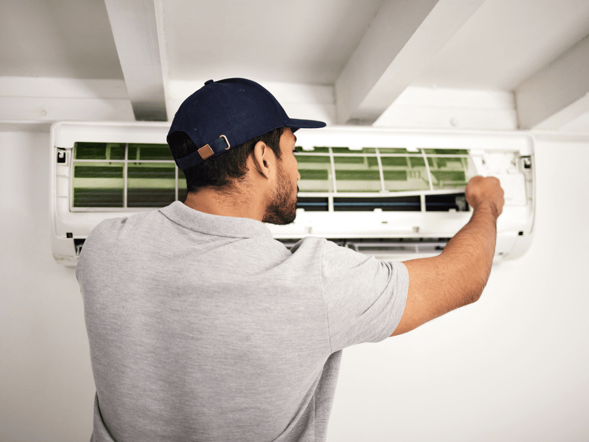 A person wearing a cap and polo shirt is inspecting or repairing a wall-mounted air conditioning unit indoors, specializing in ductless mini-split repair.