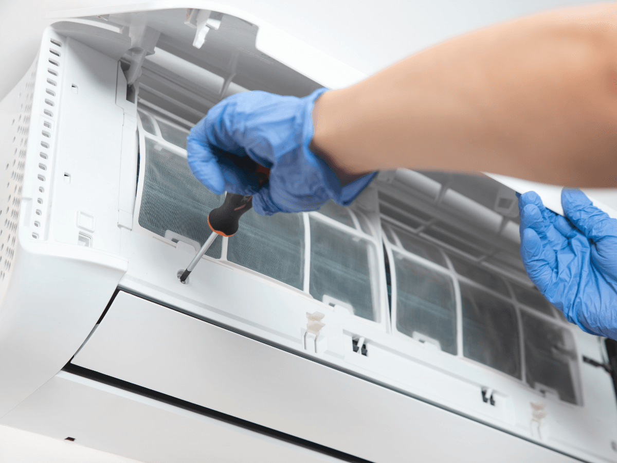 Person wearing blue gloves uses a screwdriver to repair or maintain the inside of a wall-mounted air conditioner unit, providing expert ductless mini-split repair.